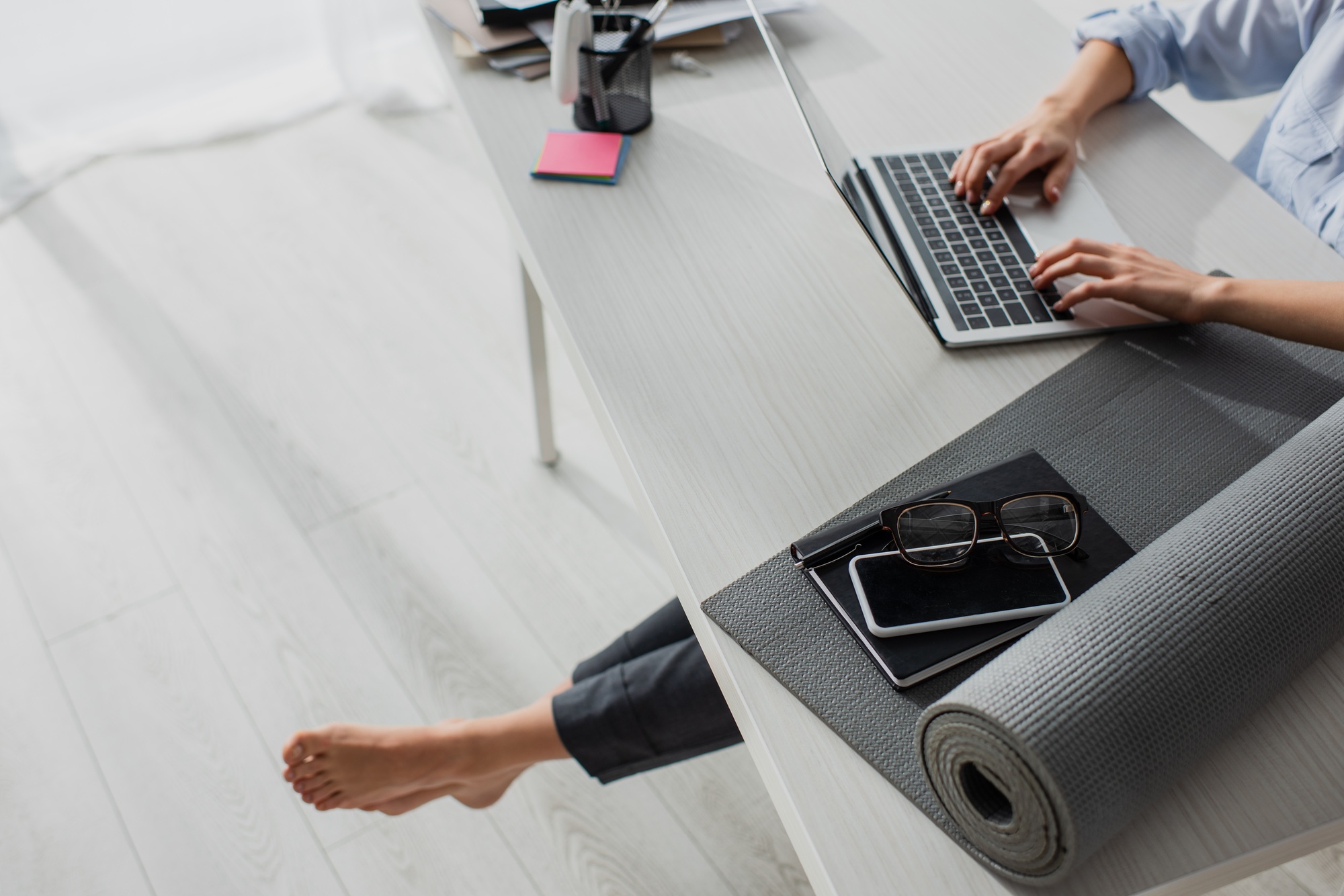 cropped view of barefoot businesswoman working on laptop at workplace with yoga mat, notepad,